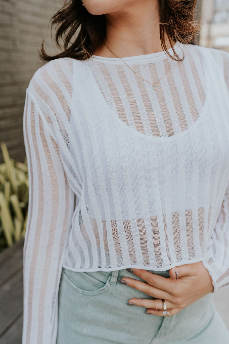 Close front view of model wearing the Dalia White Open Knit Cropped Long Sleeve Top that has white open knit fabric, a monochrome stripe pattern, a round neck, a cropped waist, and long sleeves.