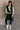 Full body front view of model wearing the Camryn Black Longline Puffer Vest features black puffer fabric, midi length, pockets, a hidden zipper with snap closures, a high neck, and a hood with drawstring ties.