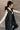 Side view of model wearing the Camryn Black Longline Puffer Vest features black puffer fabric, midi length, pockets, a hidden zipper with snap closures, a high neck, and a hood with drawstring ties.