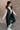Full body side view of model wearing the Camryn Black Longline Puffer Vest features black puffer fabric, midi length, pockets, a hidden zipper with snap closures, a high neck, and a hood with drawstring ties.