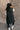 Full body back view of model wearing the Camryn Black Longline Puffer Vest features black puffer fabric, midi length, pockets, a hidden zipper with snap closures, a high neck, and a hood with drawstring ties.