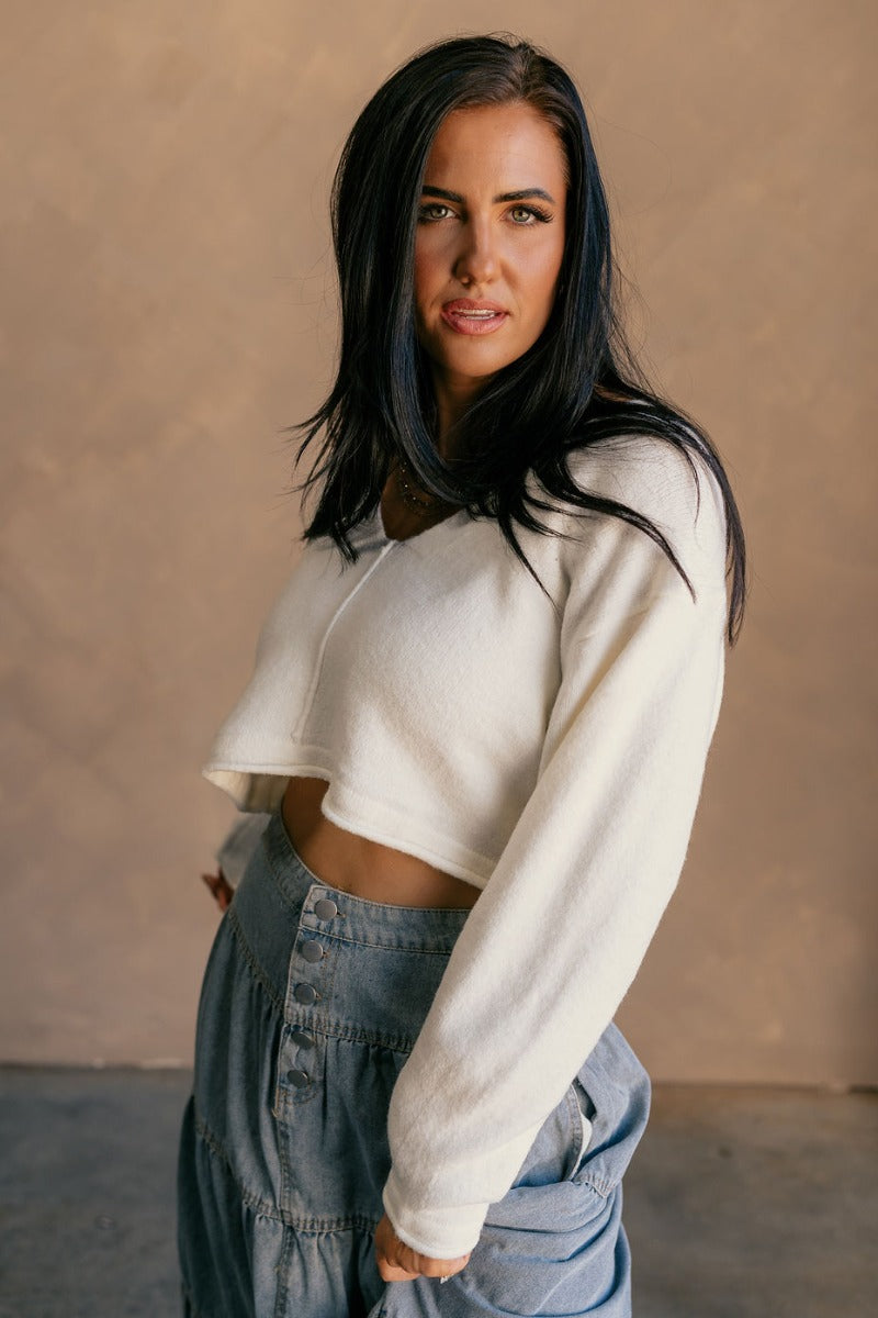 Front view of model wearing the Angela Ivory Knit long Sleeve Sweater which features off white knit fabric, a cropped waist, a v-neckline, and long sleeves.