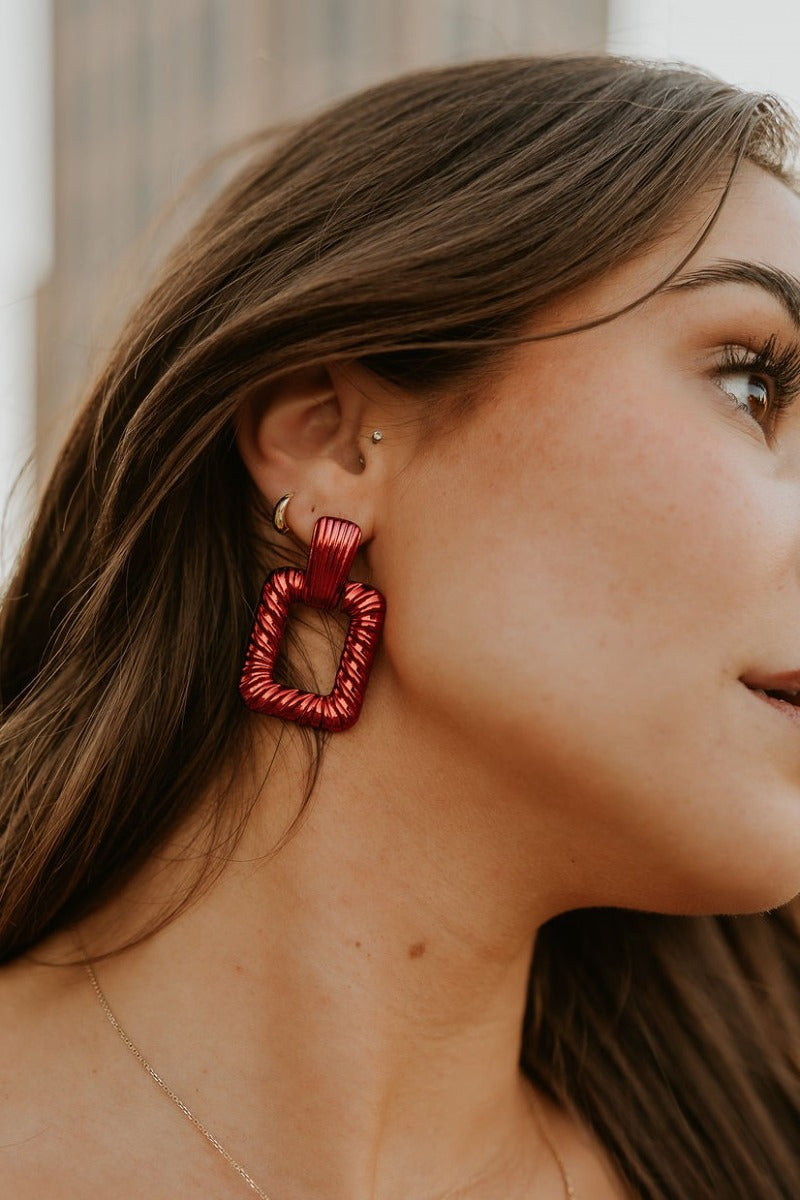 Side view of model wearing the Alina Red Earrings that have red metallic roped open rectangles with studs attached.