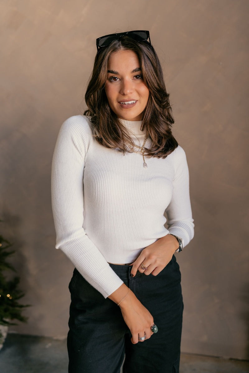 Front view of model wearing the Mila Ivory Ribbed Long Sleeve Top which features ivory ribbed knit fabric, a high neckline, and long sleeves.