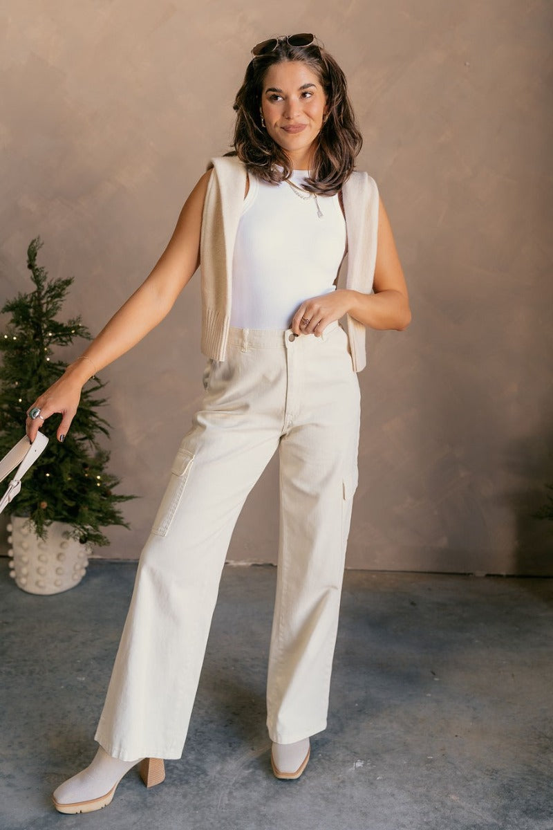 Full body front view of model wearing the Margo Cream Denim Cargo Pants that have cream cotton fabric, pockets, a front zipper with a button closure, belt loops, and wide legs.