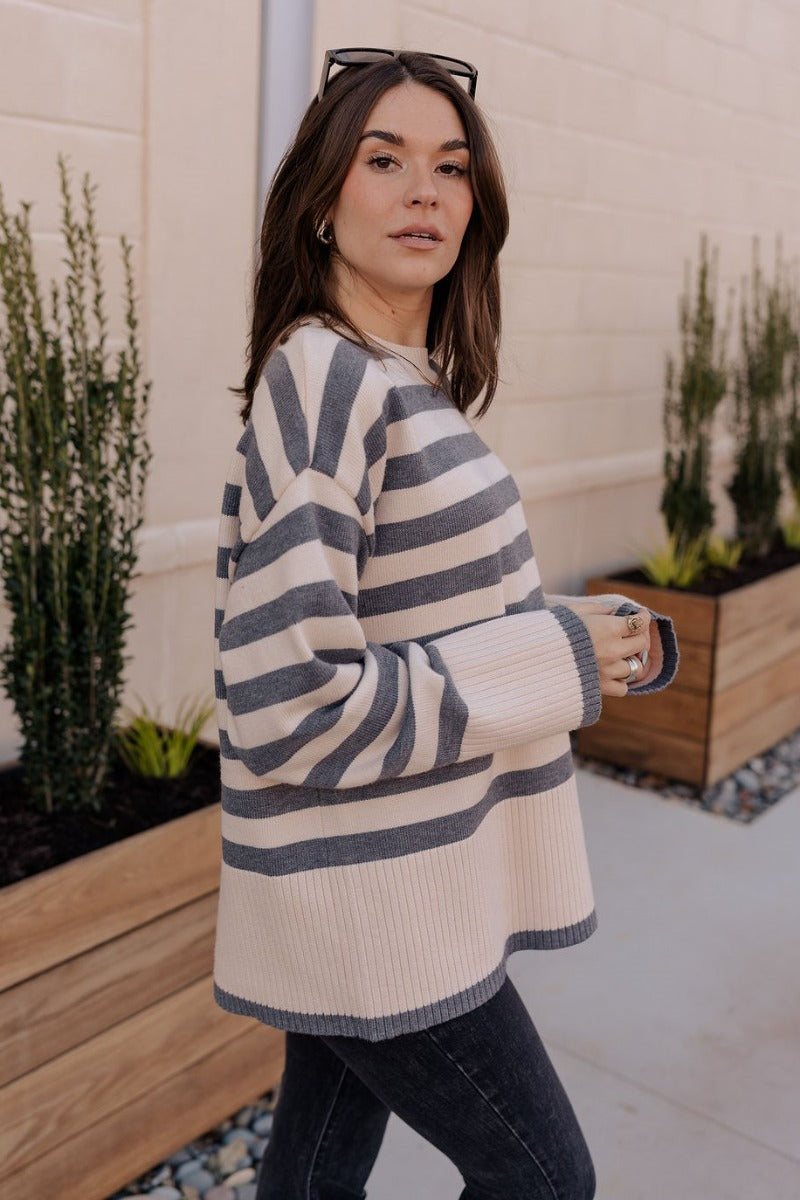 Side view of model wearing the Kelly Cream & Grey Striped Long Sleeve Sweater that has cream and grey knit fabric with a striped pattern, a ribbed hem, a round neckline, and long sleeves with ribbed cuffs.