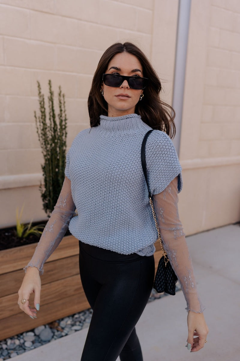 Side view of model wearing the Ryann Baby Blue Sleeveless Sweater that has light blue cable knit fabric, ribbed hem, a high neckline and a sleeveless design.