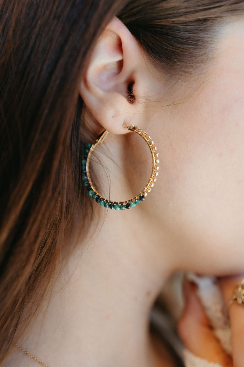 Close view of model wearing the Under The Sea Earrings that have light green and teal beads with clear stones, set in a gold hoop.