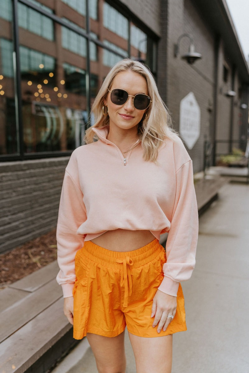 Front view of model wearing the Coastal Breeze Pullover that has peach colored fabric, a cropped waist, a monochromatic quarter zip-up, a high neckline, and long sleeves with cuffs