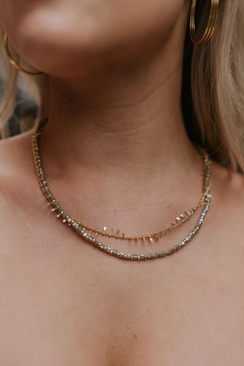 Close up image of model wearing the Should've Known Necklace that has a double gold chain with iridescent grey and gold beads.