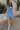 Full body view of model wearing the Phoebe Blue One- Shoulder Smocked Mini Dress which features blue light weight fabric, mini length, blue lining, upper smocked details, ruffle details, one shoulder ruffle strap and sleeveless.