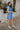 Full body side view of model wearing the Phoebe Blue One- Shoulder Smocked Mini Dress which features blue light weight fabric, mini length, blue lining, upper smocked details, ruffle details, one shoulder ruffle strap and sleeveless.