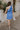Full body back view of model wearing the Phoebe Blue One- Shoulder Smocked Mini Dress which features blue light weight fabric, mini length, blue lining, upper smocked details, ruffle details, one shoulder ruffle strap and sleeveless.