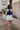 back view of model wearing The Azure White Denim Ruffle Mini Skirt features off white denim fabric, shorts lining, mini length, a ruffled skirt overlay, a flared skirt hem, and a side zipper closure.