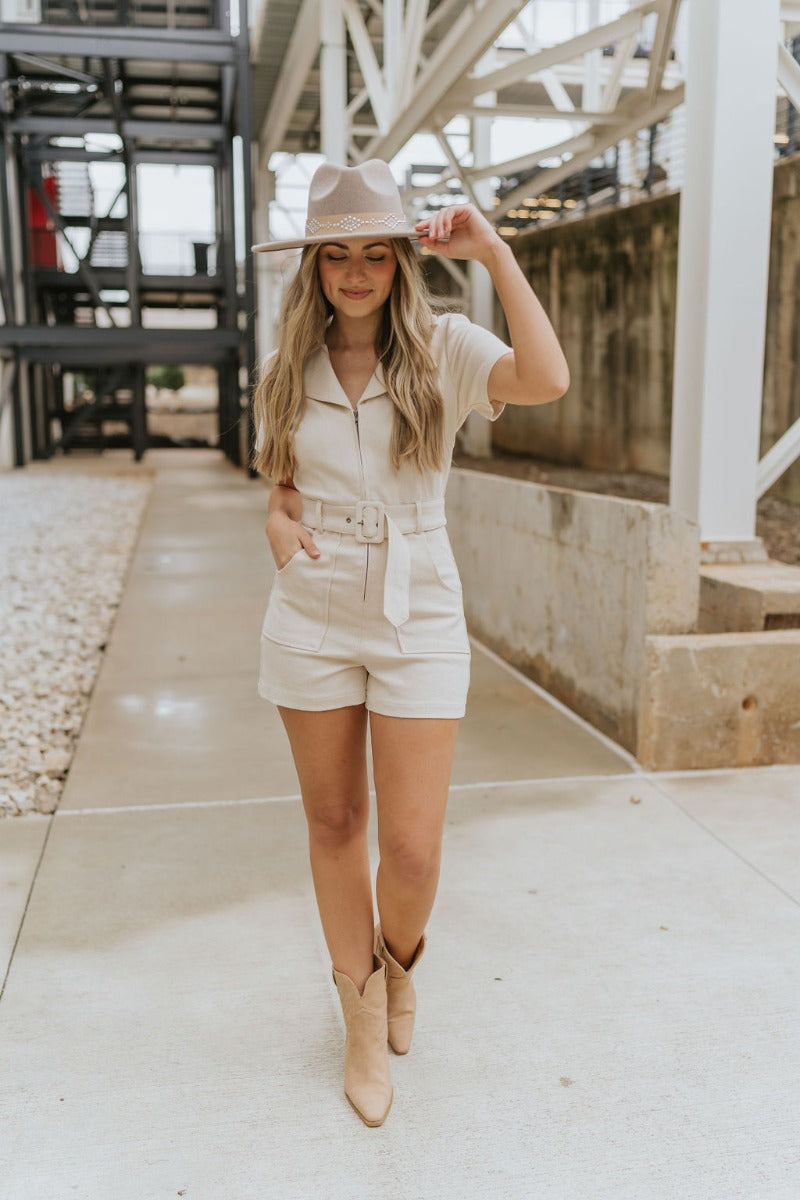 Full body front view of model wearing the Dakota Denim Romper in Oatmeal that has oatmeal denim fabric, front pockets, a front zipper, a collare, a belt at the waistline, and short sleeves
