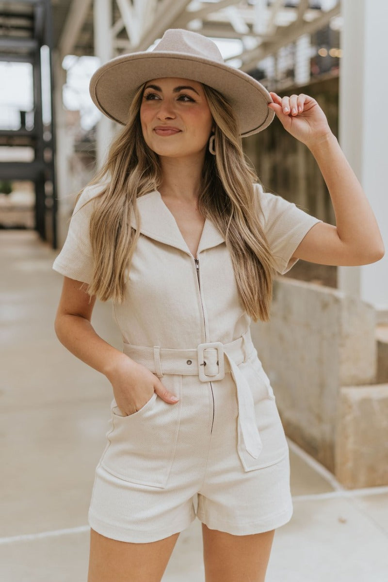 Front view of model wearing the Dakota Denim Romper in Oatmeal that has oatmeal denim fabric, front pockets, a front zipper, a collare, a belt at the waistline, and short sleeves