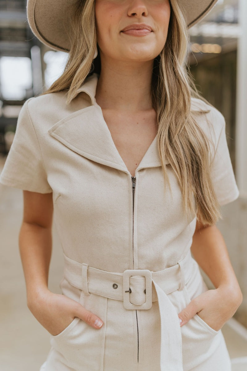 Close front view of model wearing the Dakota Denim Romper in Oatmeal that has oatmeal denim fabric, front pockets, a front zipper, a collare, a belt at the waistline, and short sleeves