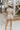 Full body back view of model wearing the Dakota Denim Romper in Oatmeal that has oatmeal denim fabric, front pockets, a front zipper, a collar, a belt at the waistline, and short sleeves
