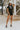 Full body front view of model wearing the Dakota Denim Romper in Black that has black denim fabric, front pockets, a hidden front zipper, a collared neckline, a monochromatic belt at the waistline, and short sleeves