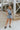 Full body front view of model wearing the Are You Ready Romper that has washed lightweight denim fabric, front pockets, two back pockets, a hidden front snap closure, a collar, short sleeves, and  frayed hems.