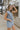 Side view of model wearing the Are You Ready Romper that has washed lightweight denim fabric, front pockets, two back pockets, a hidden front snap closure, a collar, short sleeves, and  frayed hems.