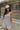 Front view of model wearing the Head In The Clouds Romper which features light grey knit fabric, pockets on each side, a quarter button-up with wooden buttons, a round neckline, and thick straps.