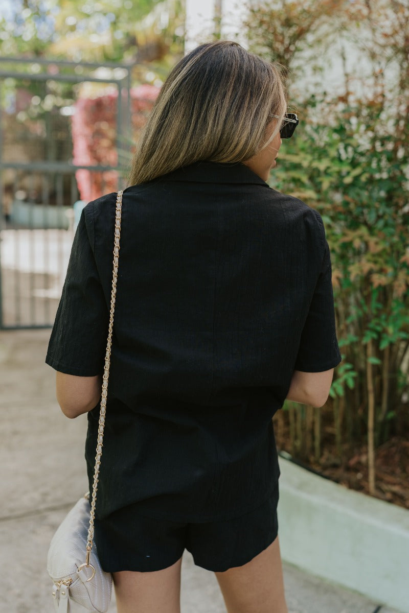 Back view of model wearing the Modern Romance Top which features black fabric, a dark tortoise front button closure, a collared neckline, and short sleeves.