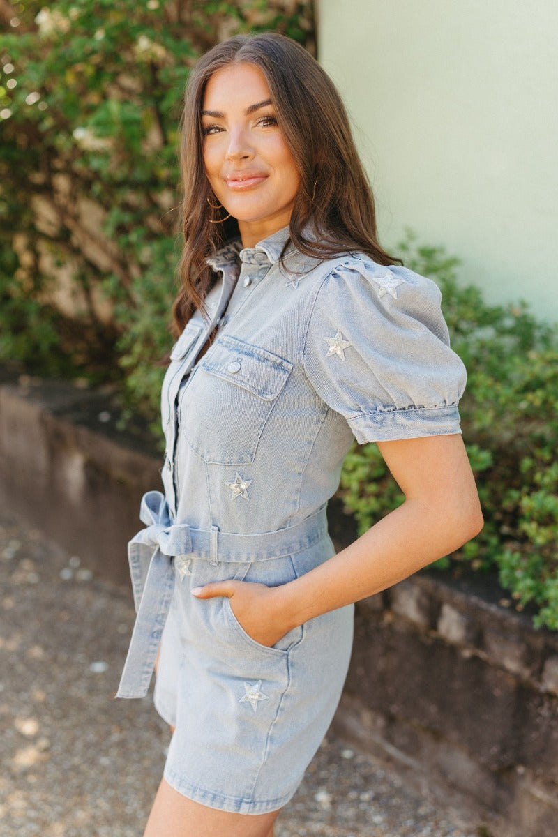 Side view of model wearing the She's A Star Denim Romper that has light denim fabric, pockets on each side, silver buttons, silver sequin star patches, a collared neckline, short puff sleeves, and denim tie belt around the waist