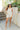 Full body front view of model wearing the Great Escape Overalls that have cream denim fabric, pockets, silver buttons, belt loops, a square neckline, adjustable straps, and distressed details
