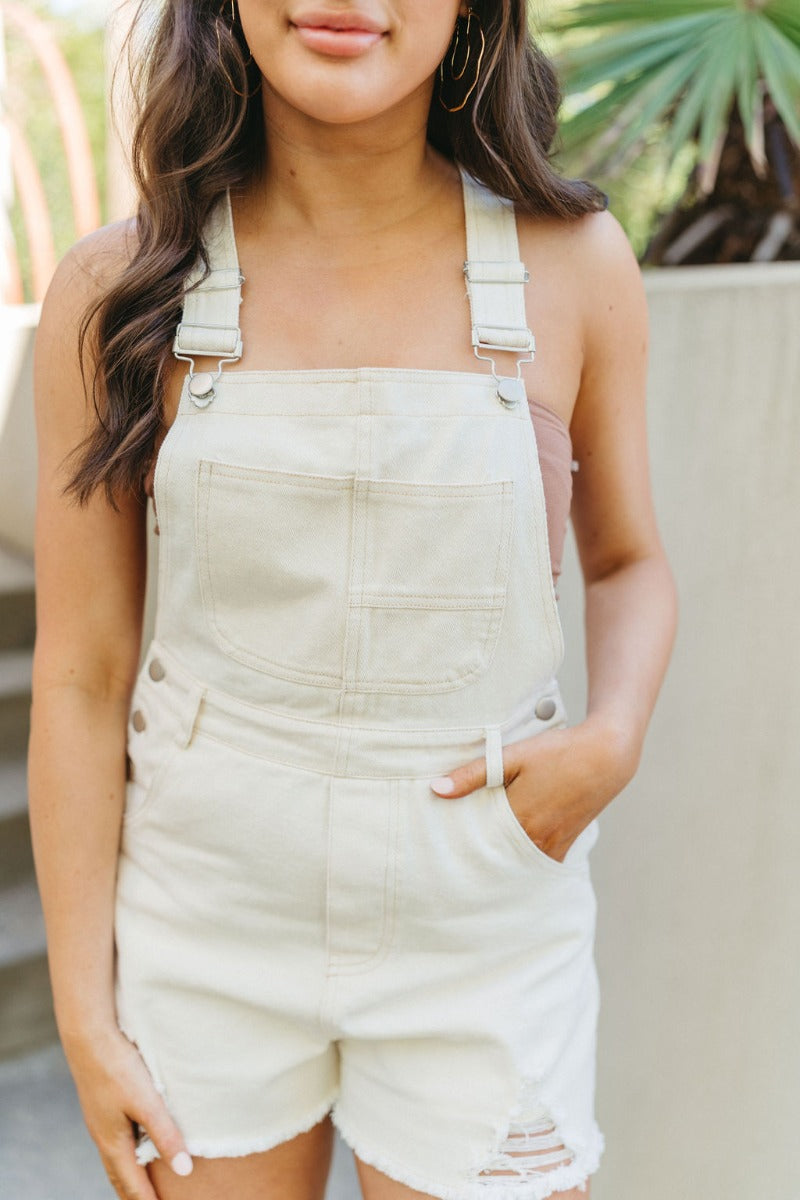 Close front view of model wearing the Great Escape Overalls that have cream denim fabric, pockets, silver buttons, belt loops, a square neckline, adjustable straps, and distressed details