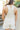 Back view of model wearing the Great Escape Overalls that have cream denim fabric, pockets, silver buttons, belt loops, a square neckline, adjustable straps, and distressed details