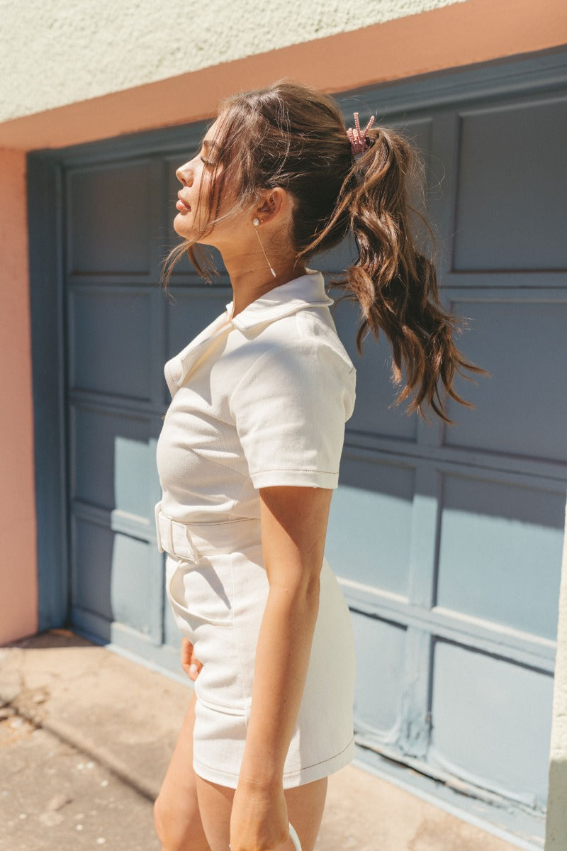 Side view of model wearing the Dakota Denim Romper in Off White which features white denim fabric, front pockets, a hidden front zipper, a collared neckline, a monochromatic belt at the waistline, and short sleeves.