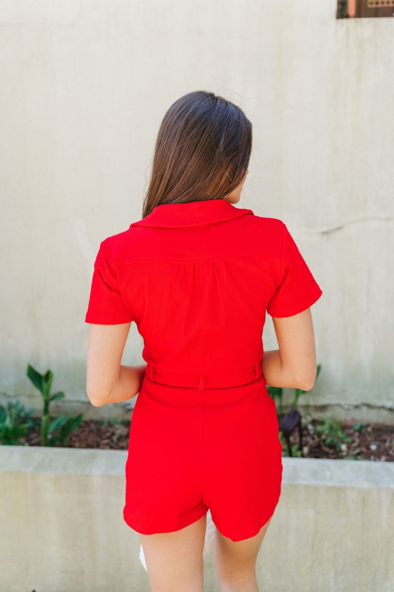 Back view of model wearing the Dakota Denim Romper in Red which features red denim fabric, front pockets, a hidden front zipper, a collared neckline, a monochromatic belt at the waistline, and short sleeves.