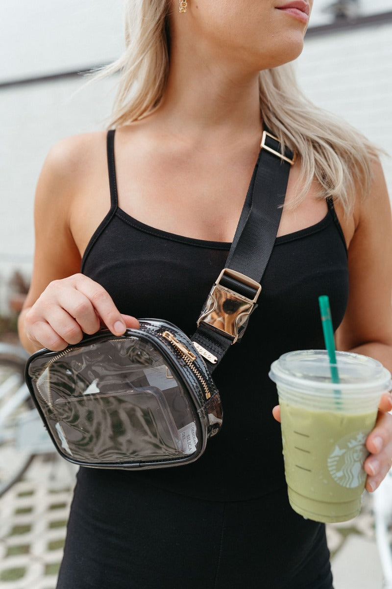 Model is wearing the Victoria Clear Sling Bag in Black that has clear fabric with black trim, a black adjustable strap with gold hardware, and a gold zipper closure.