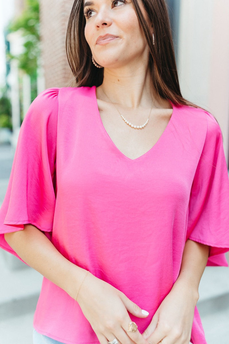 Close up view of model wearing the Better Days Top in Pink which features fuchsia satin fabric, a round low neckline, and 3/4 flare sleeves.