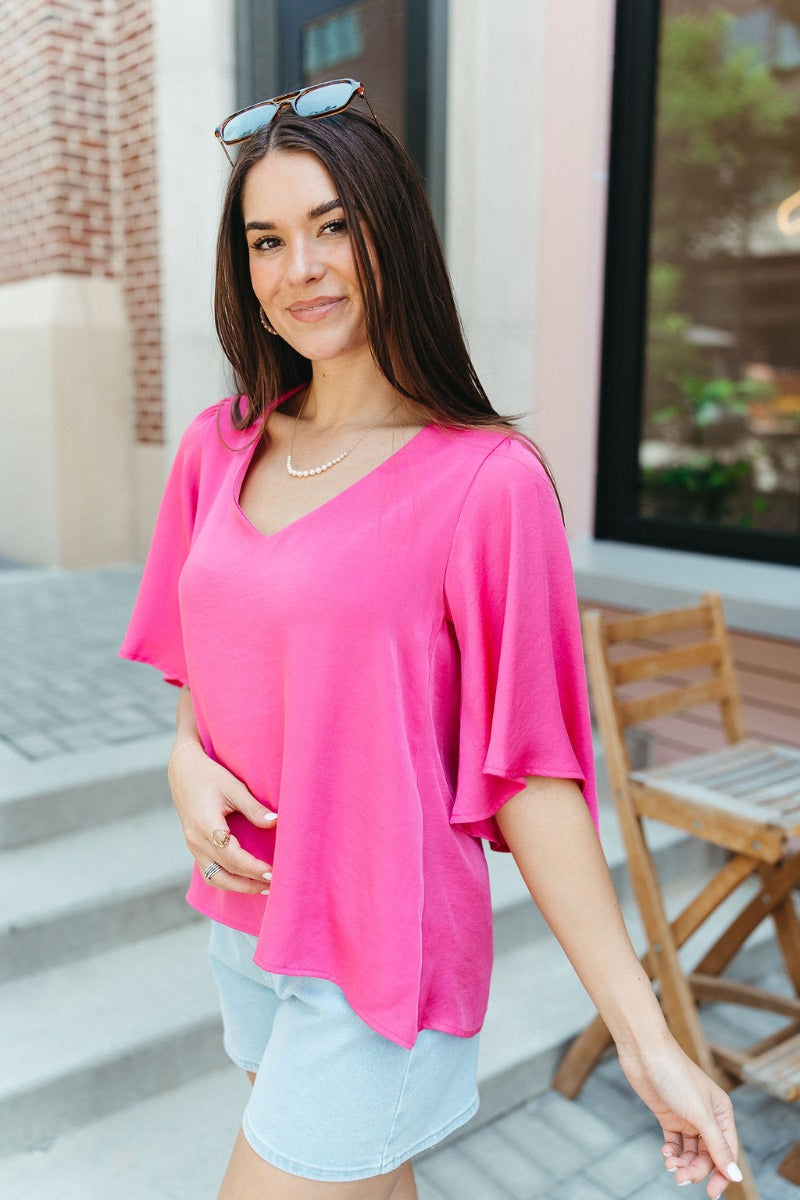 Side view of model wearing the Better Days Top in Pink which features fuchsia satin fabric, a round low neckline, and 3/4 flare sleeves.