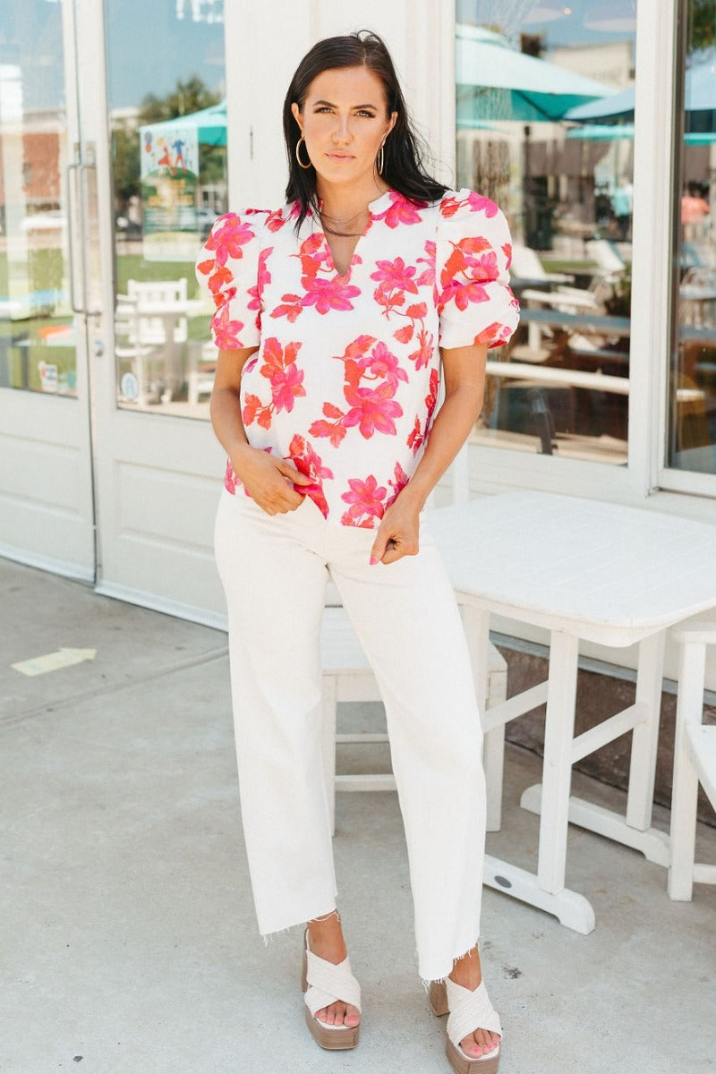 Full front of model wearing the Nothing Compares Floral Top which features white brocade fabric with a fuchsia and red floral print, white lining, round neck with a v-cutout and short puff sleeves.