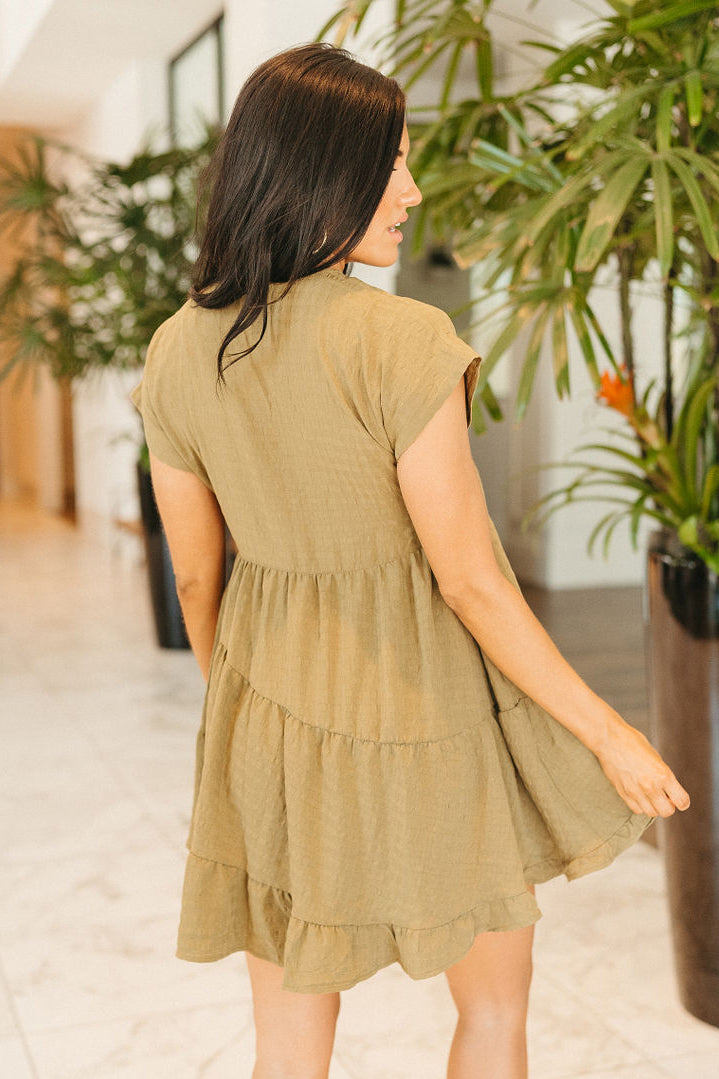 Back view of model wearing the Layla Babydoll Dress in Olive which features olive green fabric with a monochromatic checkered pattern, olive green lining, mini length, two-tiered baby doll style, a v-neckline, and short sleeves.