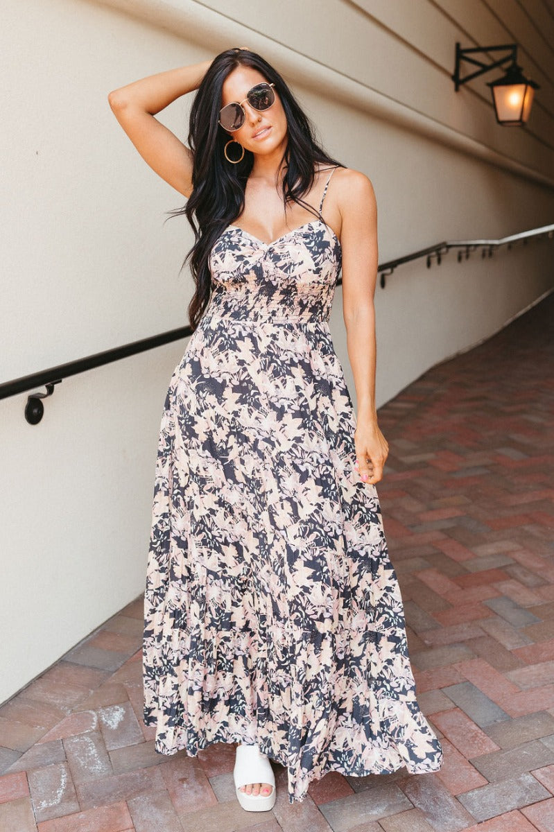 Full body view of model wearing the South Beach Floral Dress which features black fabric with a light pink, peach, lavender and tan floral pattern, a three-tiered skirt, a smocked upper, a sweetheart neckline, and adjustable straps.