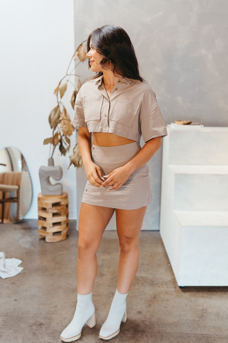 Full front view of model wearing the Next Level Crop Top that has taupe fabric, a cropped waist, front chest pockets, a monochromatic button-up front, a collared neckline and short sleeves