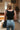 Back view of model wearing the Ready To Go Bodysuit in Black that has black knit fabric, a square neckline, a sleeveless body, and a thong bottom with snap closures