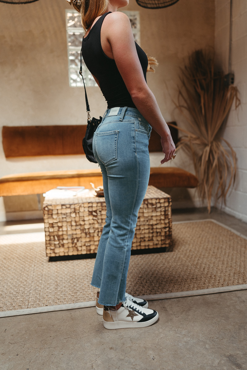 Side view of model wearing theThe Ceros: Meet Me Downtown Jeans which features medium blue denim wash, front zipper with button closure, mid-rise, two front pockets, two back pockets, belt loops and cropped flare with distressed hem.
