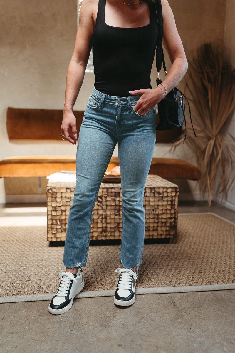 Front view of model wearing theThe Ceros: Meet Me Downtown Jeans which features medium blue denim wash, front zipper with button closure, mid-rise, two front pockets, two back pockets, belt loops and cropped flare with distressed hem.