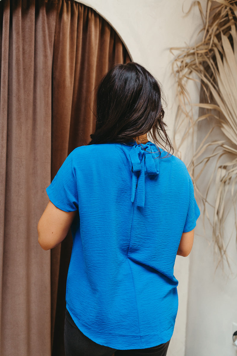 Back view of model wearing the Somebody Like You Top in Blue that has royal blue fabric, a high neckline, short sleeves and a back tie closure.