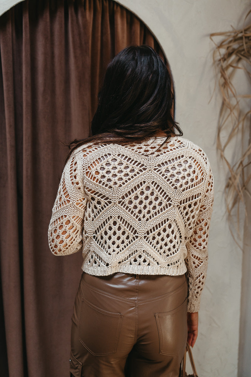 Back view of model wearing the Verona Crochet Top that has natural open-knit fabric, a cropped waist with a tie closure, and long sleeves.
