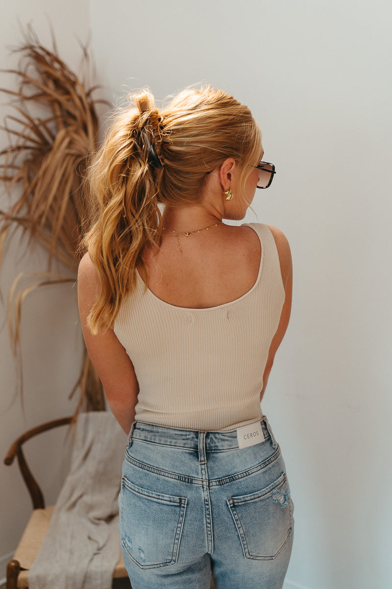 Back view of model wearing the Let's Go Ribbed Bodysuit that has oatmeal ribbed knit fabric, a scooped neckline, a sleeveless body, and a thong bottom with adjustable snap closures