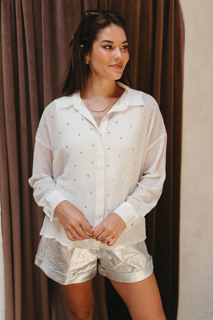 Front view of model wearing the Struck By You Top which features white sheer fabric, monochromatic buttons, a collared neckline, a rhinestone design, and long sleeves with buttoned cuffs.