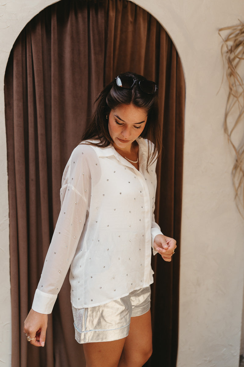 Side view of model wearing the Struck By You Top which features white sheer fabric, monochromatic buttons, a collared neckline, a rhinestone design, and long sleeves with buttoned cuffs.