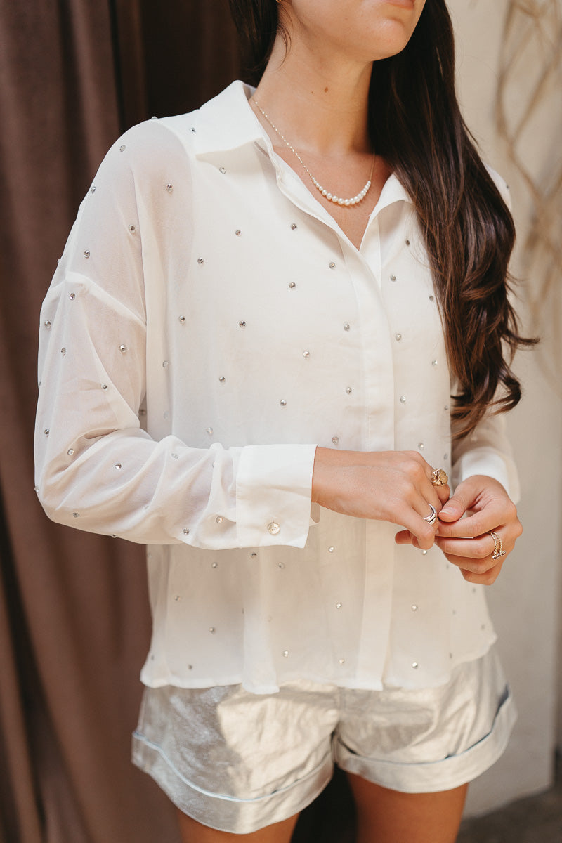 Close up view of model wearing the Struck By You Top which features white sheer fabric, monochromatic buttons, a collared neckline, a rhinestone design, and long sleeves with buttoned cuffs.