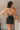Back view of model wearing the Spin Me Around Romper which features washed black denim, two front pockets, a sweetheart neckline, adjustable straps, an open back with corset ties, and a back zipper.